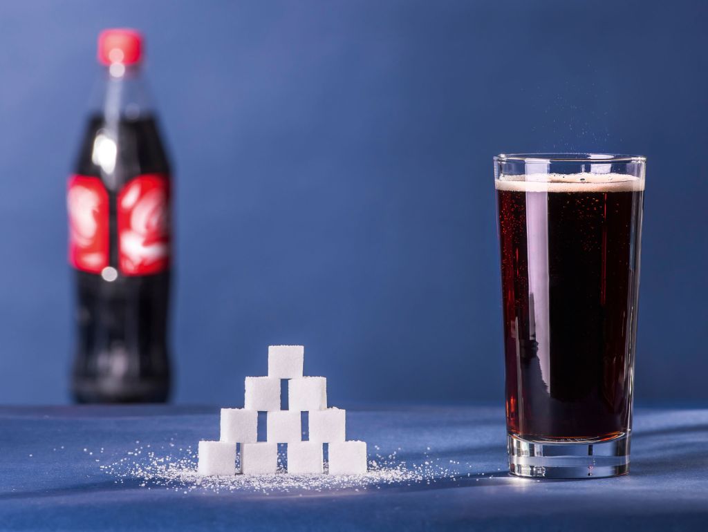 Ein Glas Cola (250 ml) enthält 30 g Zucker. Diese Menge ist in Zuckerwürfeln neben dem Glas aufgestapelt